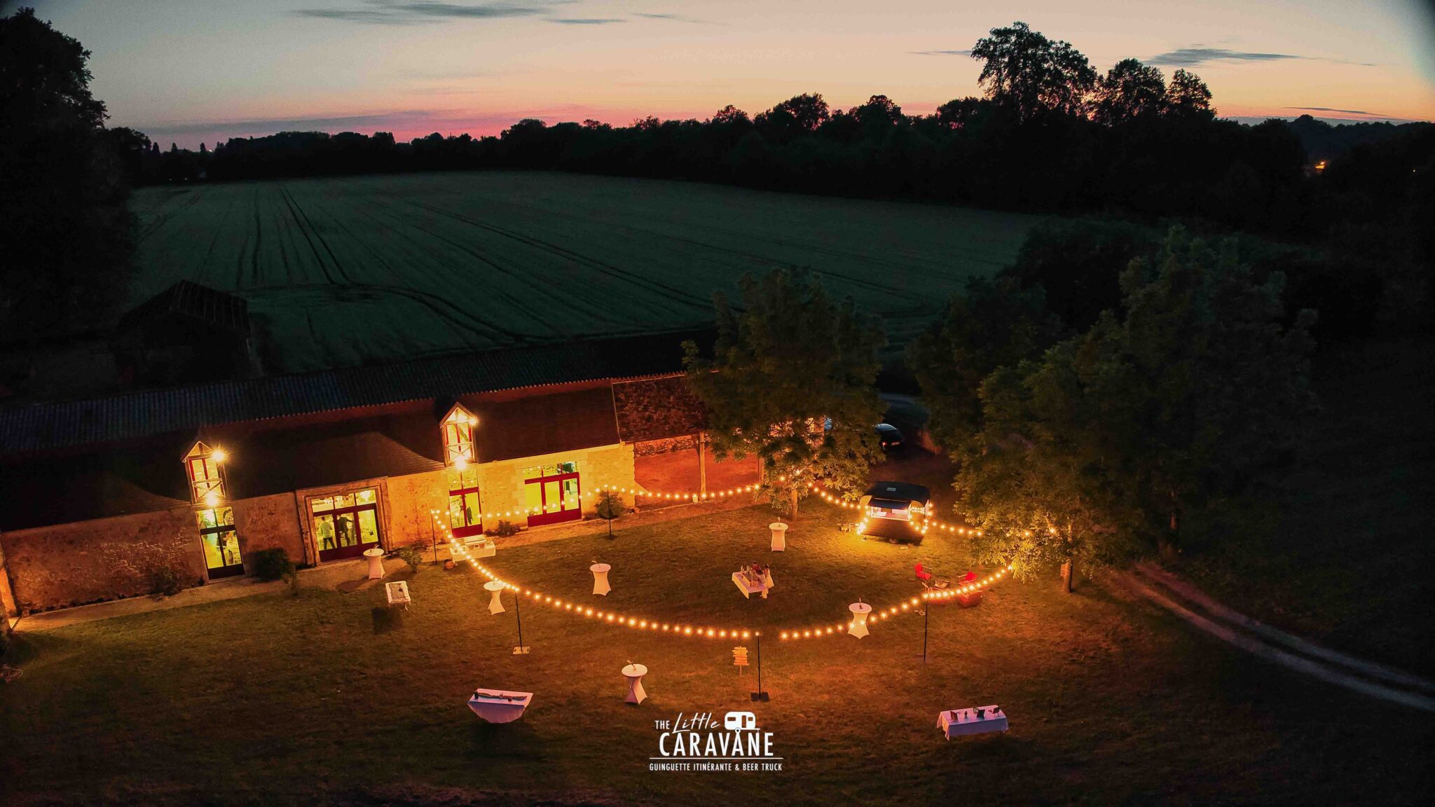 Ambiance de soirée mariage avec guirlandes lumineuses et caravane vintage en fond, bar à vinyles et bar à bières en pleine animation.