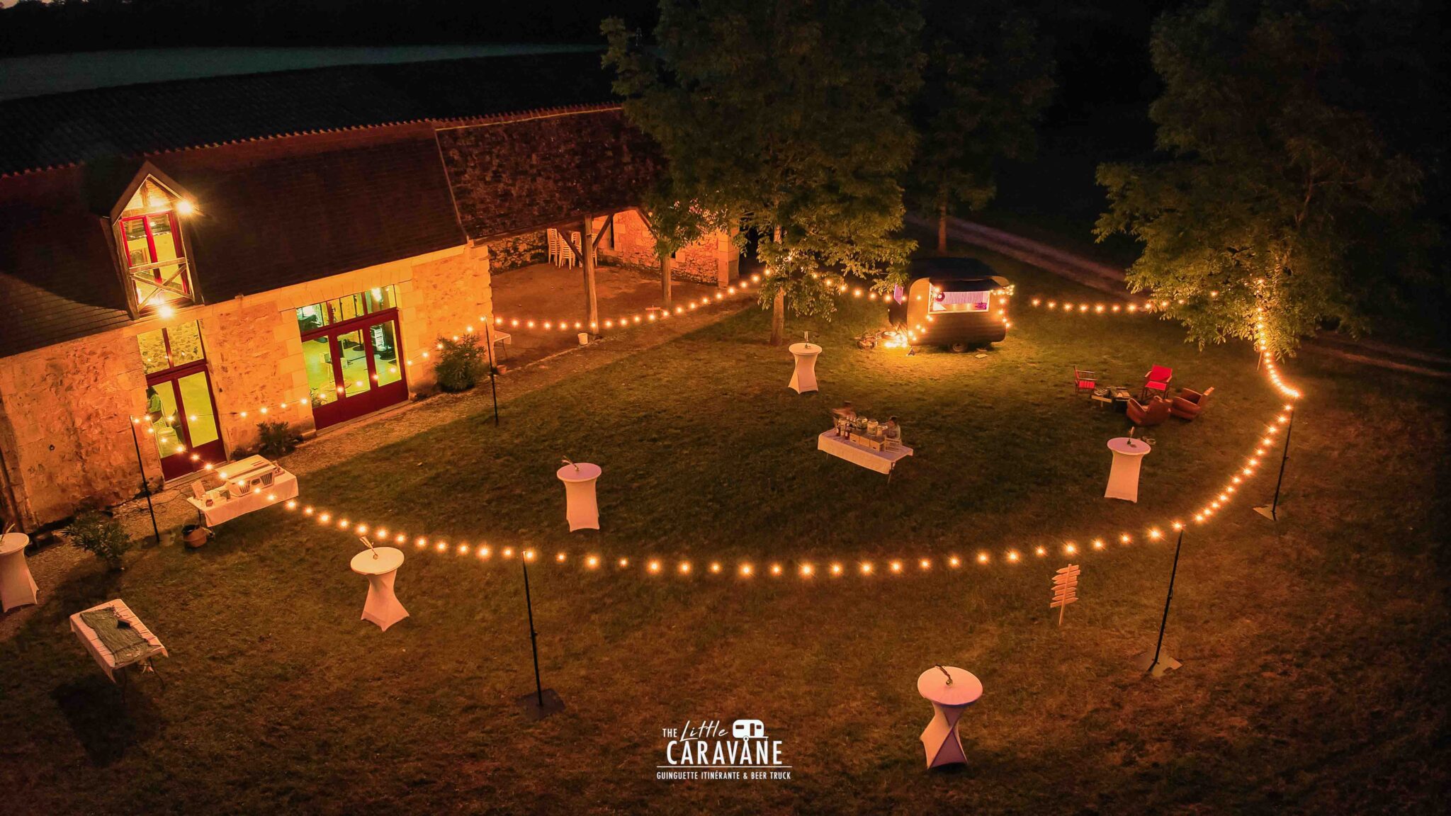 Ambiance de soirée mariage avec guirlandes lumineuses et caravane vintage en fond, bar à vinyles et bar à bières en pleine animation.
