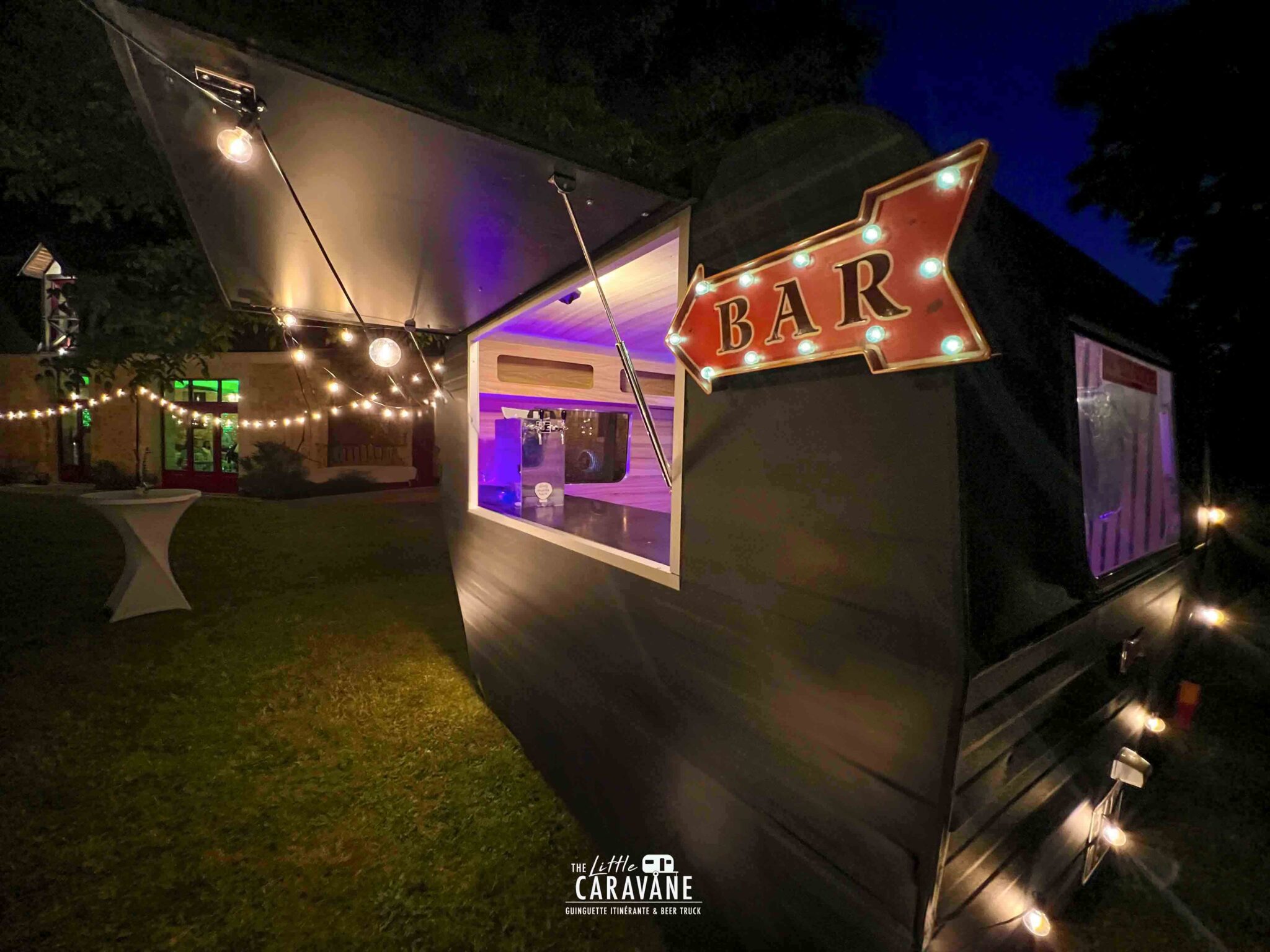 Ambiance de soirée mariage avec guirlandes lumineuses et caravane vintage en fond, bar à vinyles et bar à bières en pleine animation.